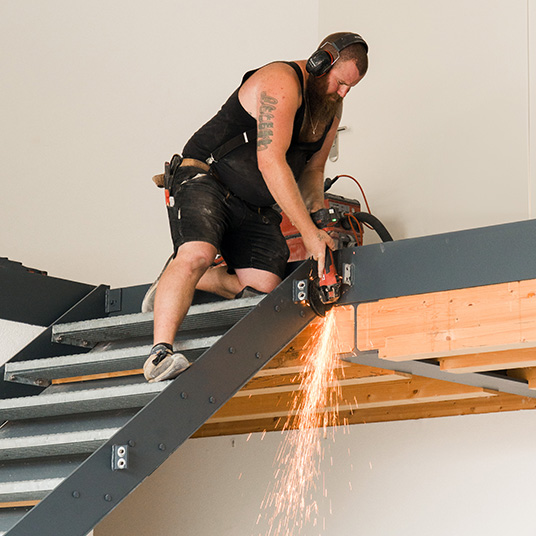 Ettwein-Mitarbeiter bearbeitet einen Stahlträger, während Funken sprühen – präzise Handwerksarbeit im modernen Holzbau