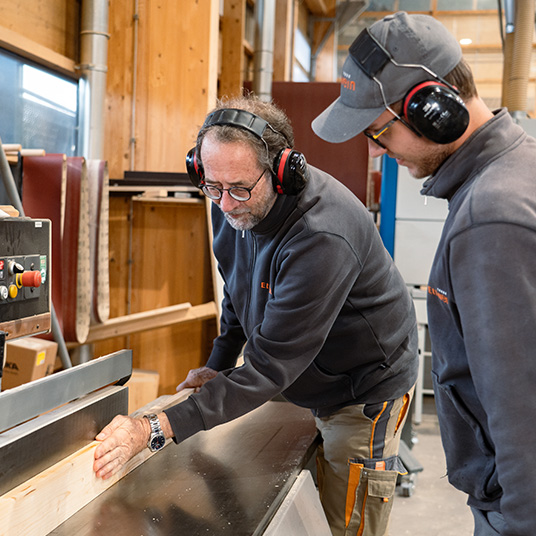 Zwei Mitarbeiter der Werner Ettwein GmbH arbeiten gemeinsam an einer Säge in der Holzfertigung und tragen Gehörschutz